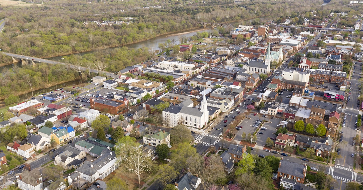 This aerial image showcases downtown Fredericksburg, Virginia, highlighting its blend of historic architecture, residential neighborhoods, and commercial areas. A river and a bridge are visible in the background, emphasizing the town's proximity to natural landmarks. The image provides context for promoting Fredericksburg as the location for RingSpaces Coworking.