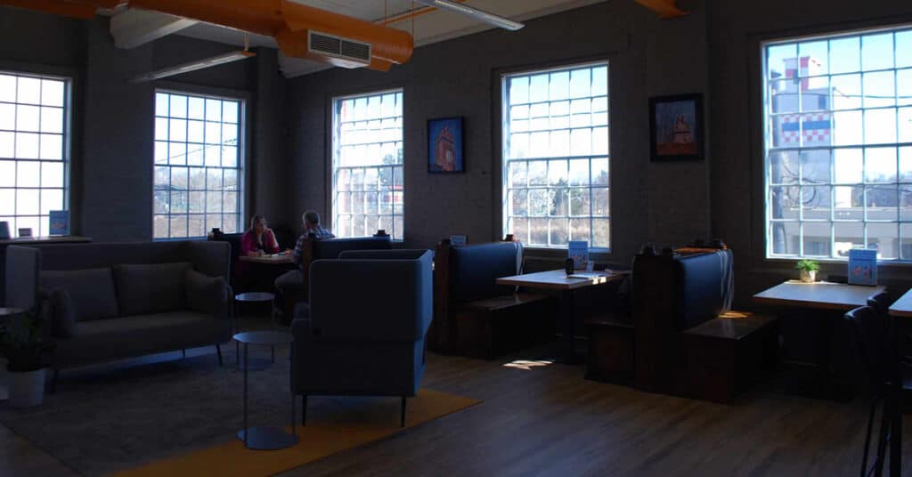 This image showcases a modern coworking and meeting space in Fredericksburg, Virginia. The interior features a mix of comfortable seating areas, including couches and booth-style tables, with large windows allowing natural light to illuminate the room. The decor is contemporary, with dark walls, bright orange ceiling accents, and framed art pieces. The space appears conducive for collaboration, relaxation, or focused work.