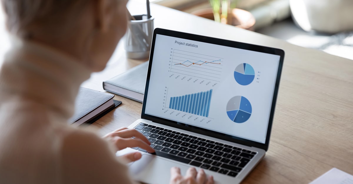 This image illustrates a freelancer analyzing project statistics on a laptop, with charts and graphs displaying progress and data insights. The setting is a well-lit workspace with a clean wooden desk, books, and a cup of pens, reflecting focus and organization. It aligns with the concept of productivity and how coworking spaces provide a structured, professional environment conducive to efficient work.