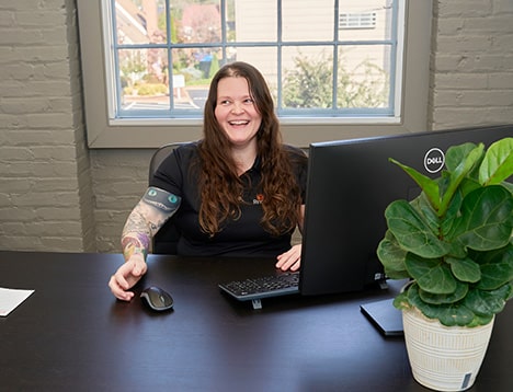 The image portrays a cheerful individual working at a desk in a private office at RingSpaces Fredericksburg. The workspace features a Dell monitor, a potted plant, and a bright window providing natural light. The casual yet professional setting highlights an environment conducive to a better work-life balance.