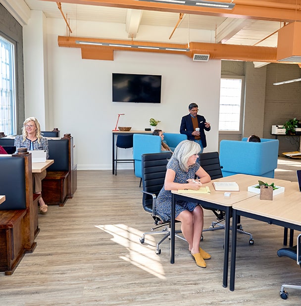 The image illustrates a dynamic coworking environment at RingSpaces Fredericksburg. It features professionals working in an open, well-lit space with a variety of seating options, including booth-style desks and collaborative tables. The space is modern, with exposed beams, a wall-mounted screen, and comfortable furnishings, fostering productivity and collaboration in a community-oriented atmosphere.