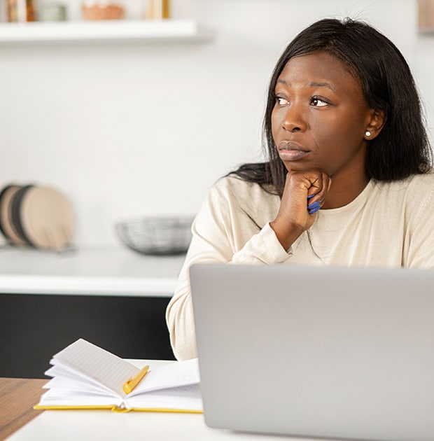 The image shows a woman sitting alone at a desk in a home setting, working with a laptop and an open notebook. She appears thoughtful, holding a pen and looking away from the screen, suggesting contemplation or problem-solving. The background includes home decor, reinforcing the idea of remote or home-based work.