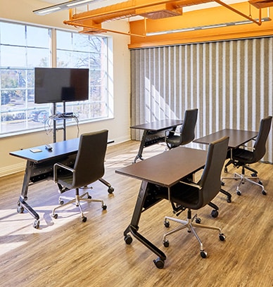 The image depicts a meeting room at RingSpaces Coworking in Fredericksburg. The room features modern furniture, including rolling desks and chairs arranged in a flexible layout, a mounted screen for presentations, and large windows providing ample natural light. The design emphasizes a professional and adaptable workspace for team meetings or training sessions.