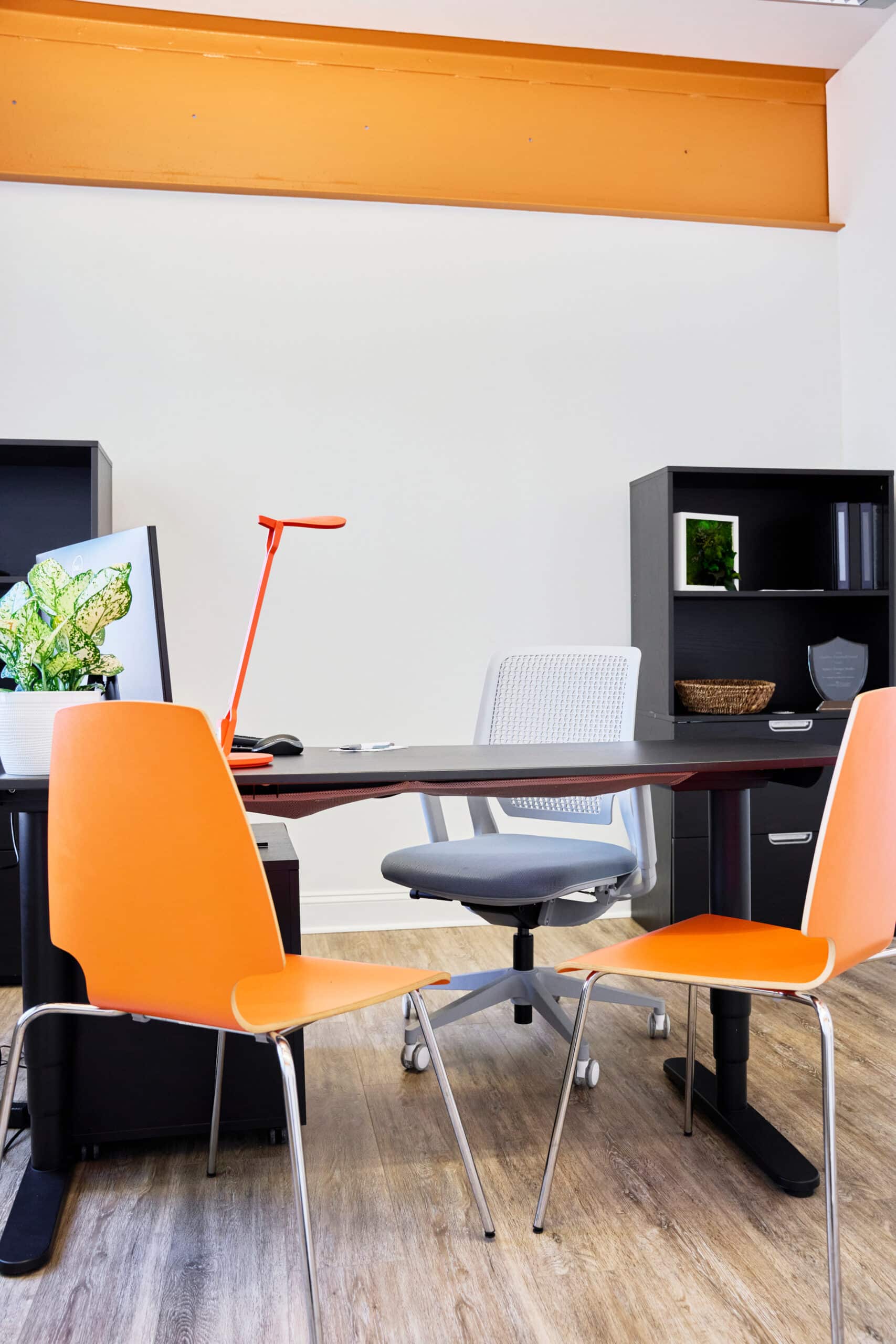 The image shows a private office setup at RingSpaces Coworking in Fredericksburg. The space features a modern design with a sleek black desk, an ergonomic office chair, and bright orange guest chairs. Additional elements include a small potted plant, a red desk lamp, and storage units with shelves and drawers, creating a functional and welcoming workspace.