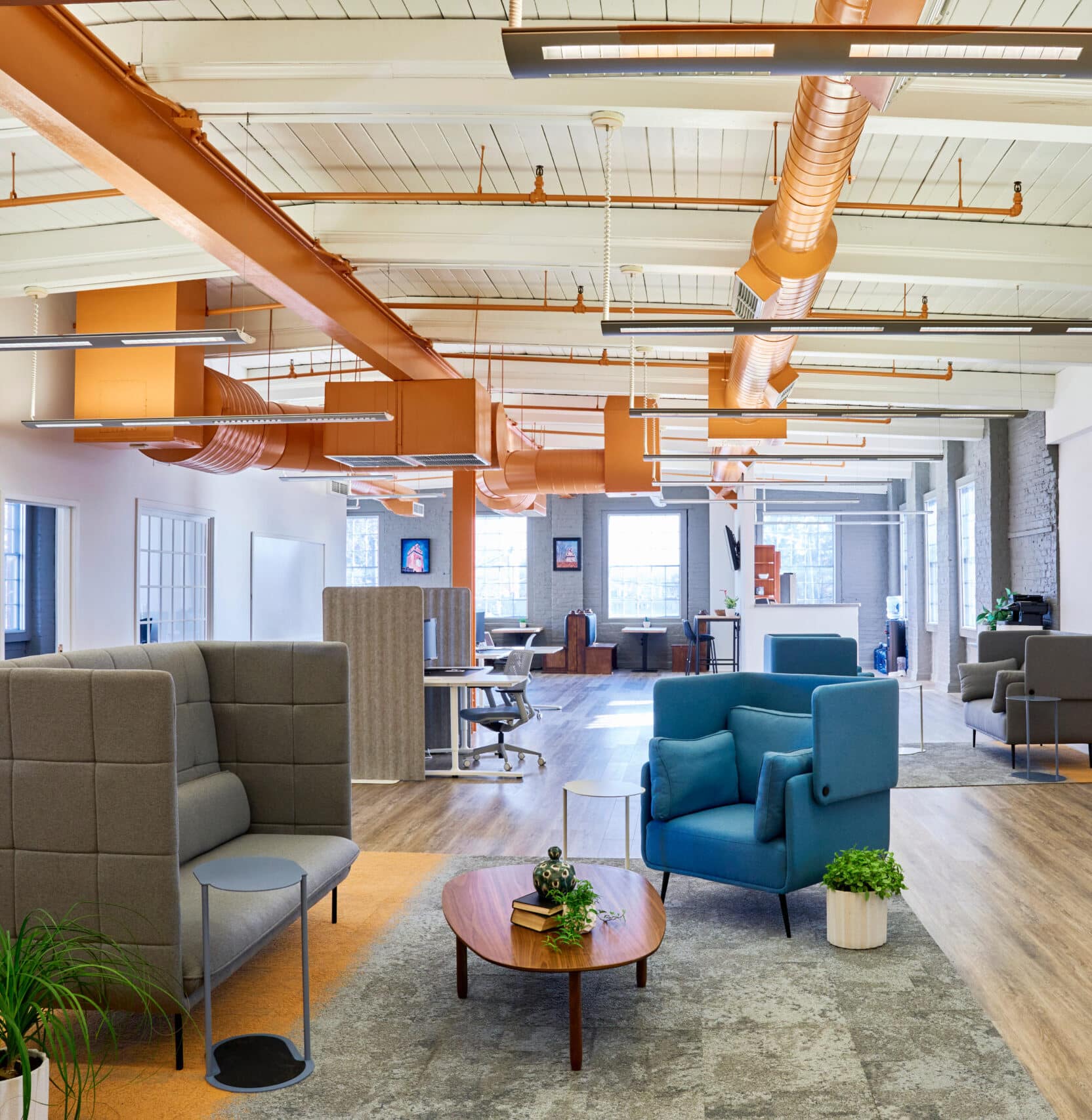 This image showcases the open-plan coworking space at RingSpaces in Fredericksburg. The interior features modern design elements with high ceilings, exposed orange ductwork, and large windows that let in natural light. The layout includes private booths, lounge seating with upholstered chairs, and communal work areas. Decor includes plants and minimalistic furniture, creating a professional yet welcoming environment. The space emphasizes collaboration and productivity while offering comfortable, designated work zones.