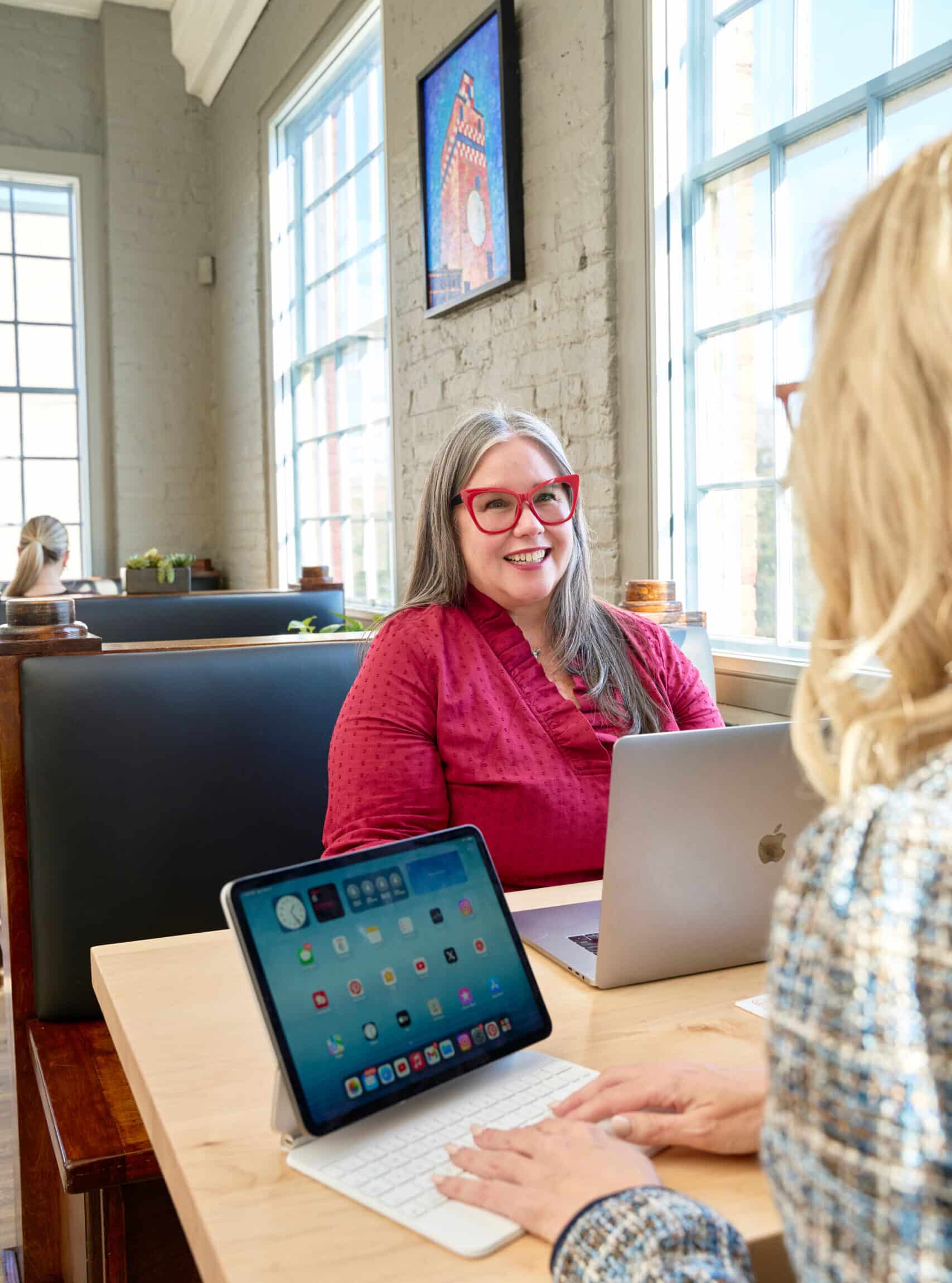 The image shows two women seated at a table in a coworking space, likely at a RingSpaces facility. One woman, wearing red-framed glasses and a pink blouse, is smiling and engaging in conversation. The other woman, partially visible, appears to be using a tablet with a keyboard case. The setting includes natural light from large windows, booth seating, and a bright, welcoming atmosphere, with artwork visible on the wall. The environment conveys a professional yet relaxed workspace.