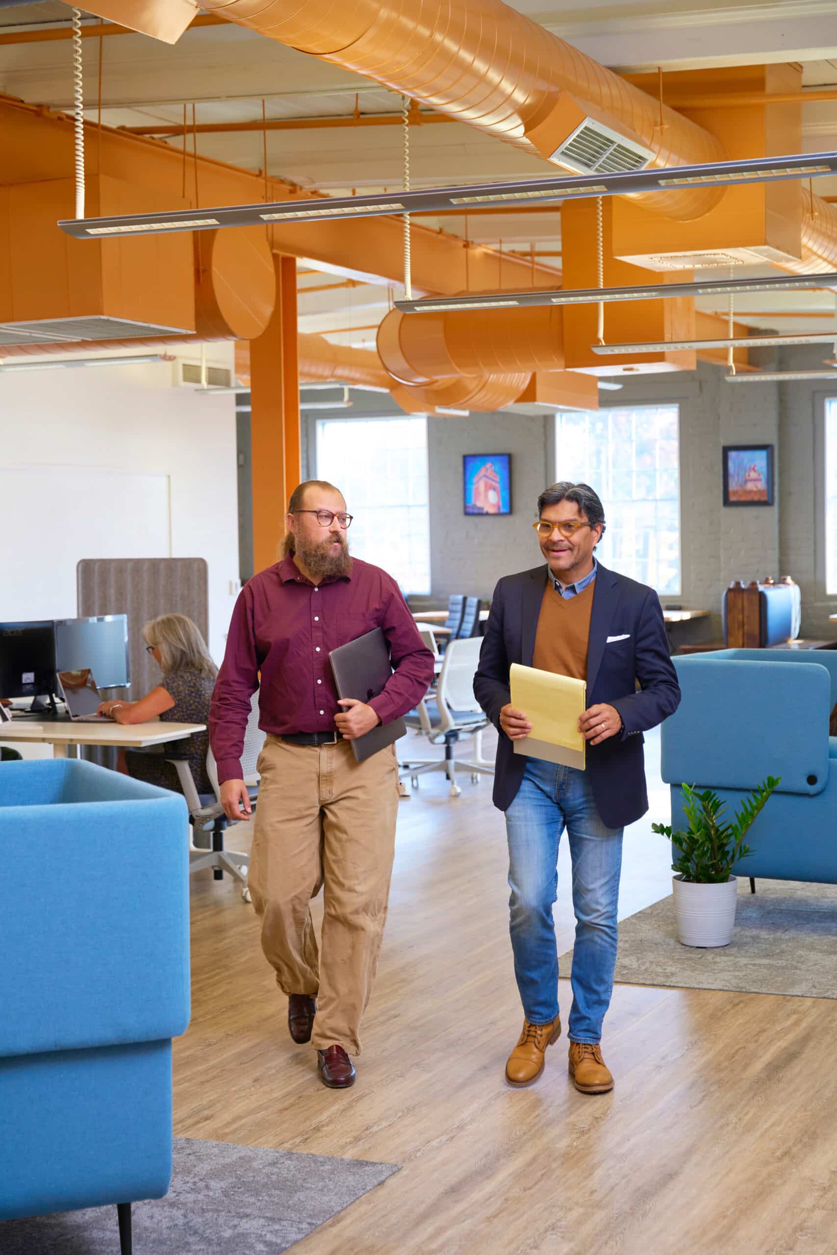 The image shows two men walking and conversing in a modern coworking space, characterized by bright natural light, vibrant orange ductwork, and a mix of open desks and private seating areas. A woman works at a computer in the background, highlighting the collaborative and productive atmosphere. This appears to be a snapshot of small business professionals engaging in a shared office environment.