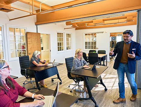 The image shows a modern office setting at RingSpaces Fredericksburg, featuring a small team meeting. A professional presenter is speaking to three participants seated at desks, each using laptops. The environment is bright, with open spaces, glass-paned walls, and exposed beams, creating a collaborative and professional atmosphere. The setting emphasizes coworking and team collaboration.