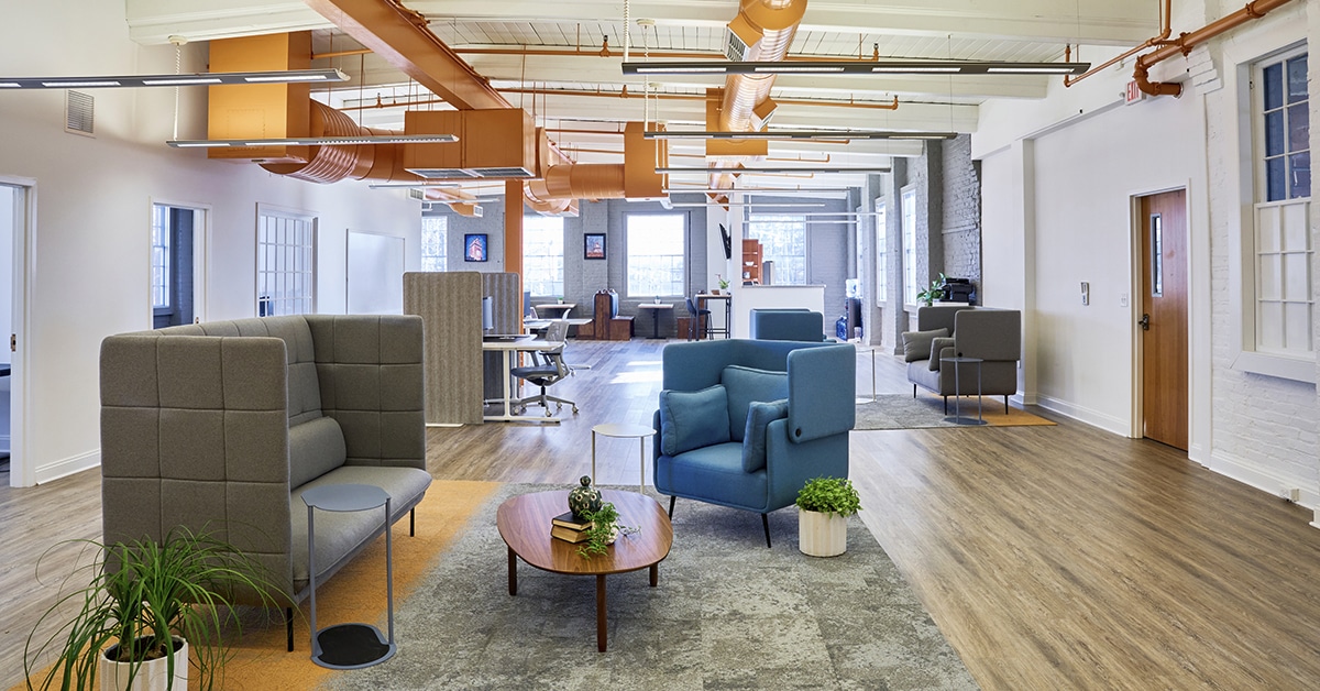 This image showcases a vibrant and modern coworking space with an open layout. It features comfortable seating areas, private booths, and collaborative workstations, promoting flexibility and productivity. The space is well-lit with large windows, contemporary furniture, and plants, creating an inviting atmosphere. Bright orange ducts and wooden floors add warmth and character, exemplifying an innovative environment ideal for coworking.