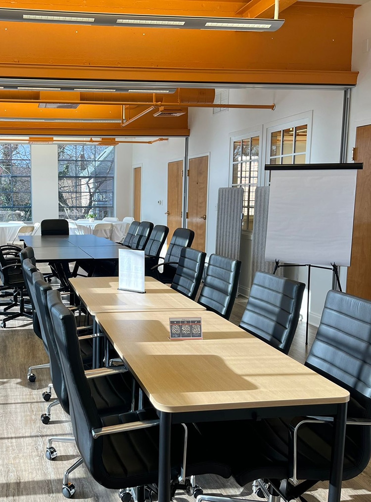 The image showcases a professional meeting space at RingSpaces of Fredericksburg, ideal for hosting business events and meetings. The room features long wooden tables, comfortable black leather chairs, and ample seating for attendees. A flip chart and large windows allowing natural light enhance the functionality and ambiance of the space. The bright orange ceiling accents add a modern and vibrant touch, reinforcing the venue’s suitability for productive gatherings.