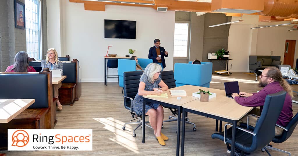 The image illustrates a dynamic coworking environment at RingSpaces Fredericksburg. It features professionals working in an open, well-lit space with a variety of seating options, including booth-style desks and collaborative tables. The space is modern, with exposed beams, a wall-mounted screen, and comfortable furnishings, fostering productivity and collaboration in a community-oriented atmosphere.