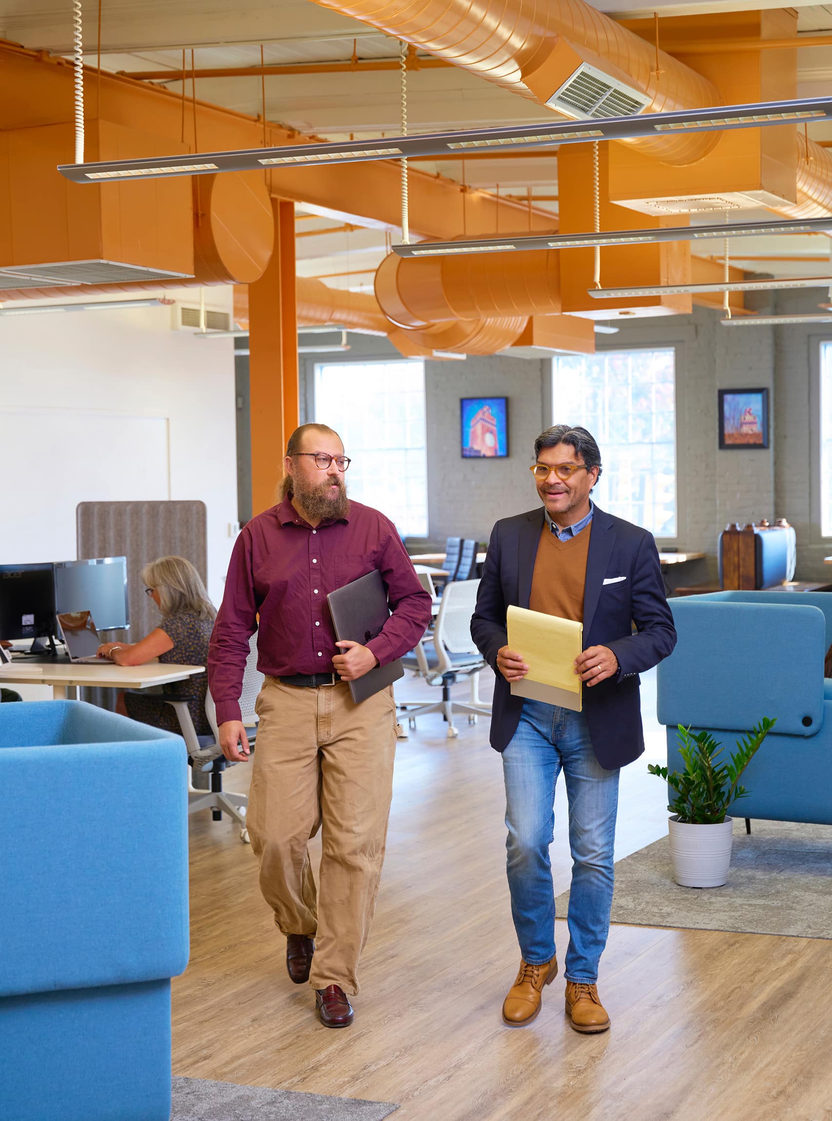 The image depicts two men walking through a modern, open-concept office space. One man is holding a laptop and the other a notepad. The office features bright orange overhead ductwork, blue office furniture, and natural light streaming through large windows. Other workers are visible in the background, seated at desks. The setting emphasizes collaboration and a contemporary workplace environment.