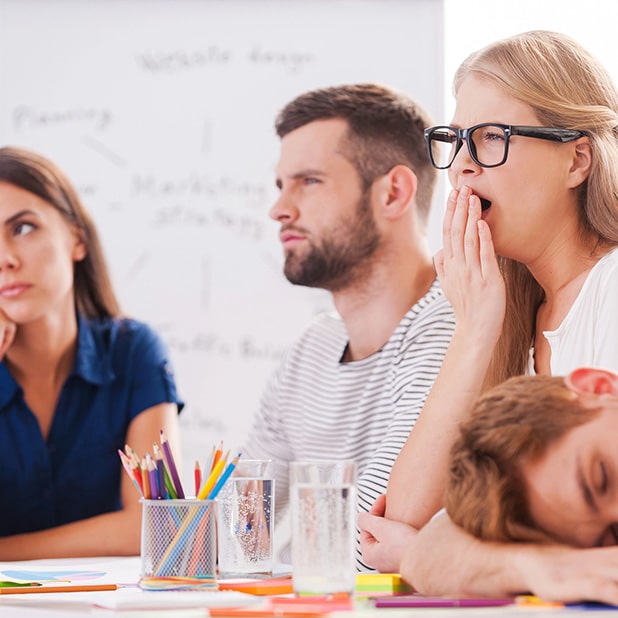 The image humorously contrasts a disengaged group in a dull meeting setting to emphasize that events at RingSpaces of Fredericksburg are anything but boring. It depicts individuals yawning and appearing uninterested, with markers and materials on the table and a whiteboard in the background. This highlights how RingSpaces provides a dynamic and engaging environment to host events that energize and inspire participants.