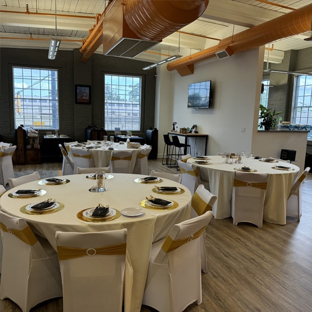 The image highlights a stylishly arranged event space featuring multiple round tables draped in white linens, each adorned with gold-accented table settings and chairs tied with gold sashes. The room has a modern industrial feel, with exposed ductwork, large windows allowing ample natural light, and sleek decor. The layout demonstrates the expertise of RingSpaces' event team in creating an inviting and professional setting for private gatherings.