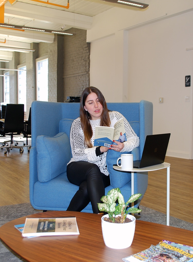 The image highlights the comfort and versatility of the coworking environment at RingSpaces of Fredericksburg. A woman is seated in a cozy blue armchair, reading a book with a laptop and coffee mug on a small side table, emphasizing flexibility and relaxation. A wooden coffee table with plants and magazines adds to the welcoming and casual ambiance, showcasing the balance of productivity and comfort available in the space.