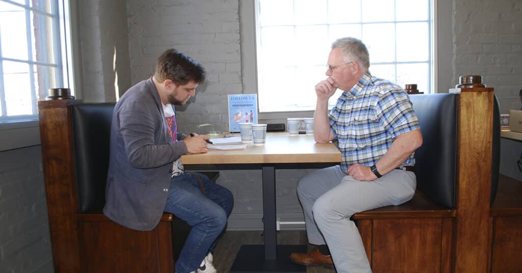 This image depicts two individuals engaged in a focused discussion at a booth in a casual, flexible workspace. The setting highlights a modern work environment with natural lighting, minimalistic decor, and a relaxed atmosphere, contrasting with a more formal traditional office space. A sign in the background suggests a social media presence, reinforcing the contemporary nature of the workspace.