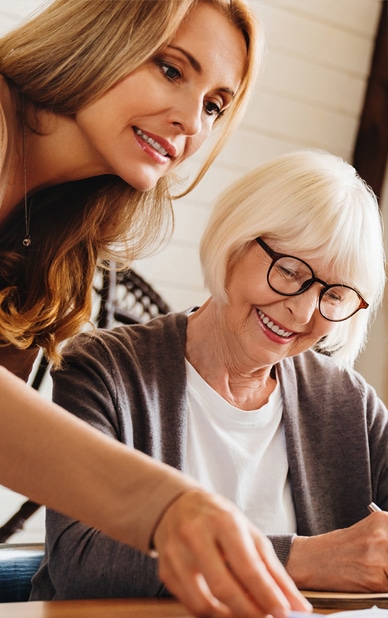 The image features a smiling woman assisting an older woman, who is engaged in writing or planning. The younger woman leans in attentively, suggesting a collaborative and supportive environment. This conveys the attentive, personalized approach of an event planner at RingSpaces of Fredericksburg, dedicated to making private events seamless and enjoyable.