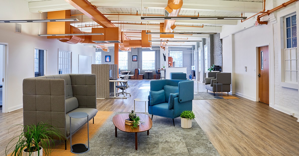 This image showcases the open-plan coworking space at RingSpaces in Fredericksburg. The interior features modern design elements with high ceilings, exposed orange ductwork, and large windows that let in natural light. The layout includes private booths, lounge seating with upholstered chairs, and communal work areas. Decor includes plants and minimalistic furniture, creating a professional yet welcoming environment. The space emphasizes collaboration and productivity while offering comfortable, designated work zones.