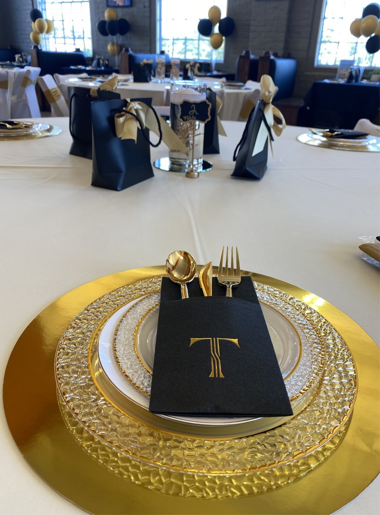 The image showcases an elegantly arranged table setting featuring gold and black accents. A black napkin with a gold "T" design holds gold utensils, placed on layered plates with textured gold trim atop a gold charger. Gift bags with gold ribbons serve as party favors, and the background includes additional decorated tables with gold and black balloons. This highlights the refined and festive ambiance at RingSpaces of Fredericksburg, perfect for private events.