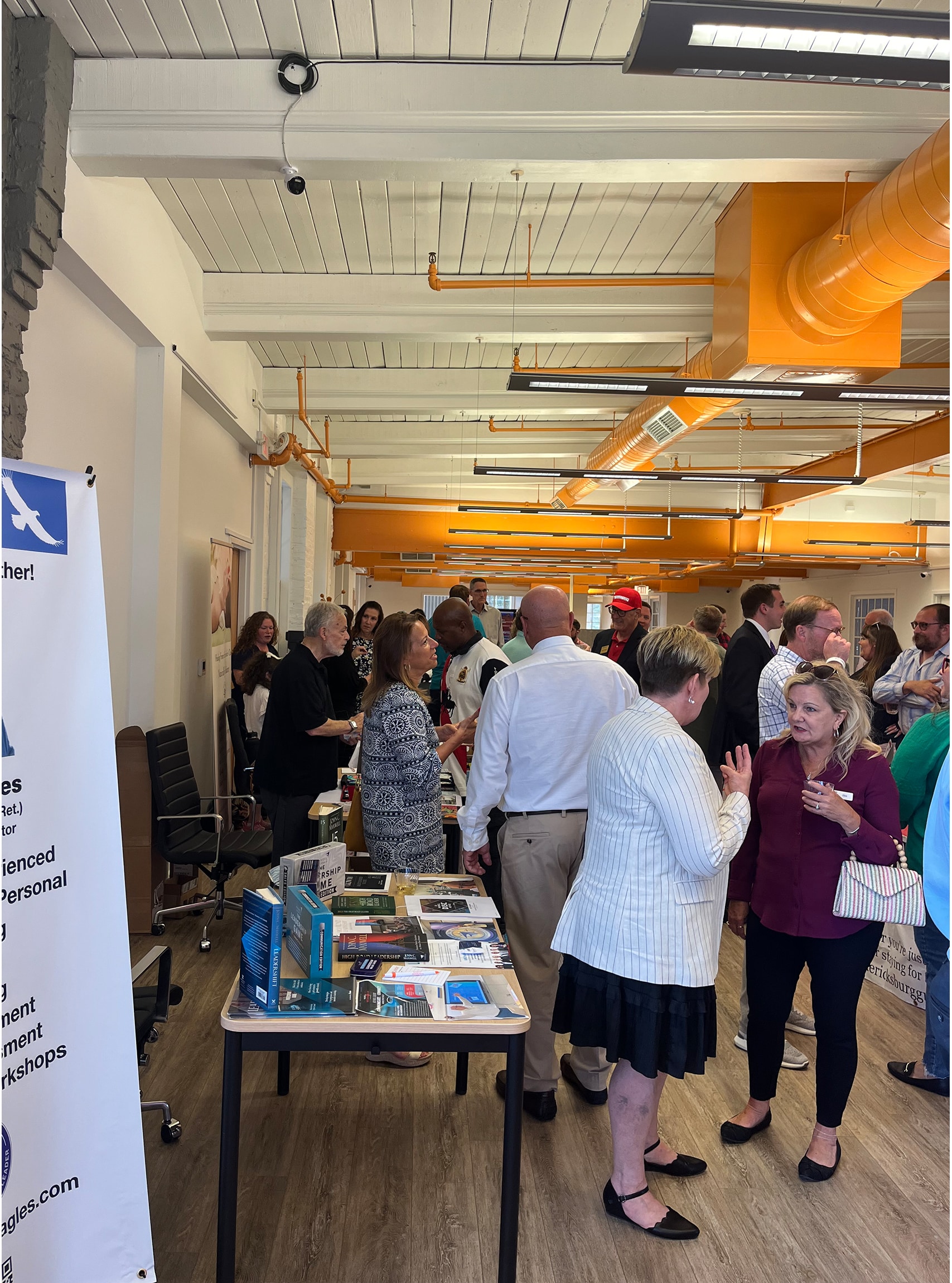 The image captures a lively business event at RingSpaces of Fredericksburg. Attendees are networking and engaging in conversations, with tables displaying books, materials, and promotional items. The open, modern coworking space, featuring bright orange ductwork and wood-style flooring, serves as a vibrant backdrop for the gathering. The event highlights RingSpaces' capability to host professional networking and business-focused occasions in a collaborative environment.