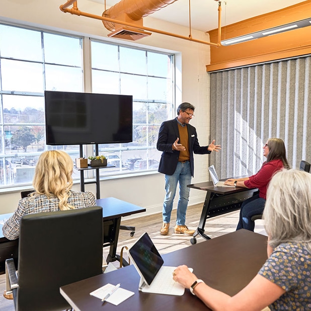 The image highlights a business training session at RingSpaces of Fredericksburg, showcasing a professional and collaborative environment. A speaker is presenting to a small group seated at desks equipped with laptops, emphasizing a focus on learning and engagement. The space features large windows providing natural light, a mounted TV for visual aids, and modern furnishings, including black chairs and wooden tables. The orange industrial ceiling elements add a vibrant touch, making it an ideal setting for productive and impactful training sessions.