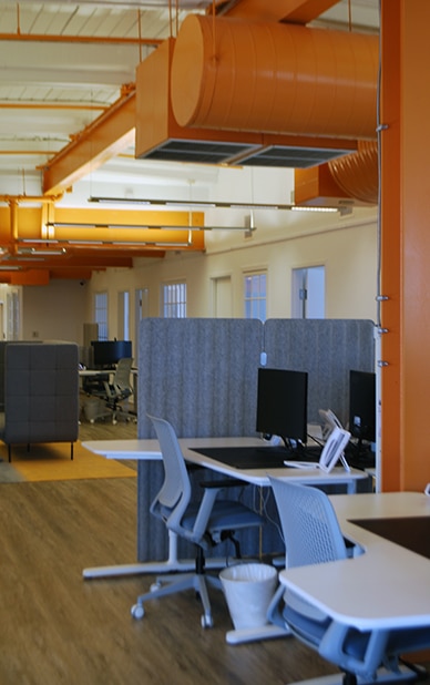 The image shows a modern coworking office space with an open-plan layout. It features individual desks equipped with ergonomic chairs, computer monitors, and dividers for privacy. The space is brightly lit and accented with orange industrial-style air ducts, creating a vibrant atmosphere. The flooring appears to be wood or laminate, adding a warm touch. This setup highlights the workspace environment available for rent at RingSpaces Coworking in Fredericksburg.