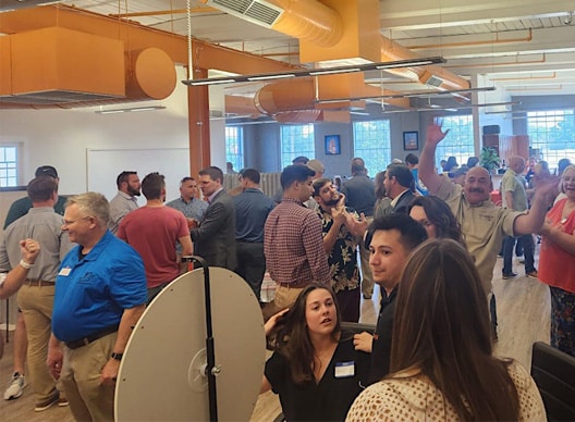 This image showcases a lively networking event at RingSpaces of Fredericksburg. The setting features a bright, open office space with modern orange ductwork and large windows. Attendees are engaged in conversations, mingling, and enjoying the event's casual, welcoming atmosphere. The image highlights RingSpaces as an ideal venue for hosting professional gatherings and networking opportunities.