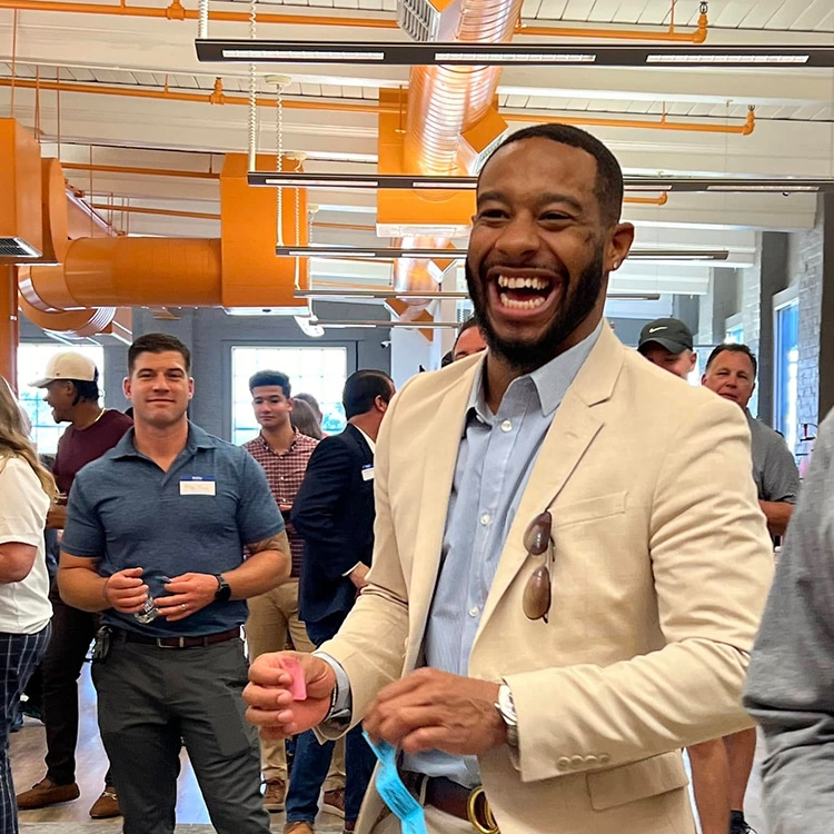 The image captures a lively moment at a business event or party hosted at RingSpaces of Fredericksburg. A smiling attendee in professional attire stands out, radiating positive energy, while others engage in conversations in the background. The setting features vibrant orange industrial ceiling accents, large windows, and a modern coworking space ambiance, emphasizing RingSpaces as a venue for creating memorable and engaging professional and social events.