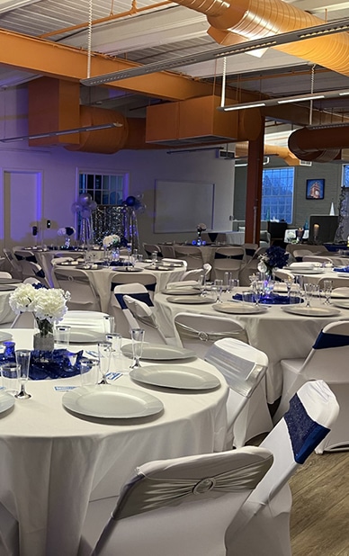 The image showcases a chic and modern event space set up for a formal gathering. Tables are dressed in crisp white linens with blue sequined centerpieces and white floral arrangements. Chairs feature white covers with blue sashes and decorative silver accents. The ambient blue lighting complements the industrial-style exposed ductwork, creating a sophisticated yet contemporary atmosphere. This highlights RingSpaces in Fredericksburg as a prime location for private event rentals.