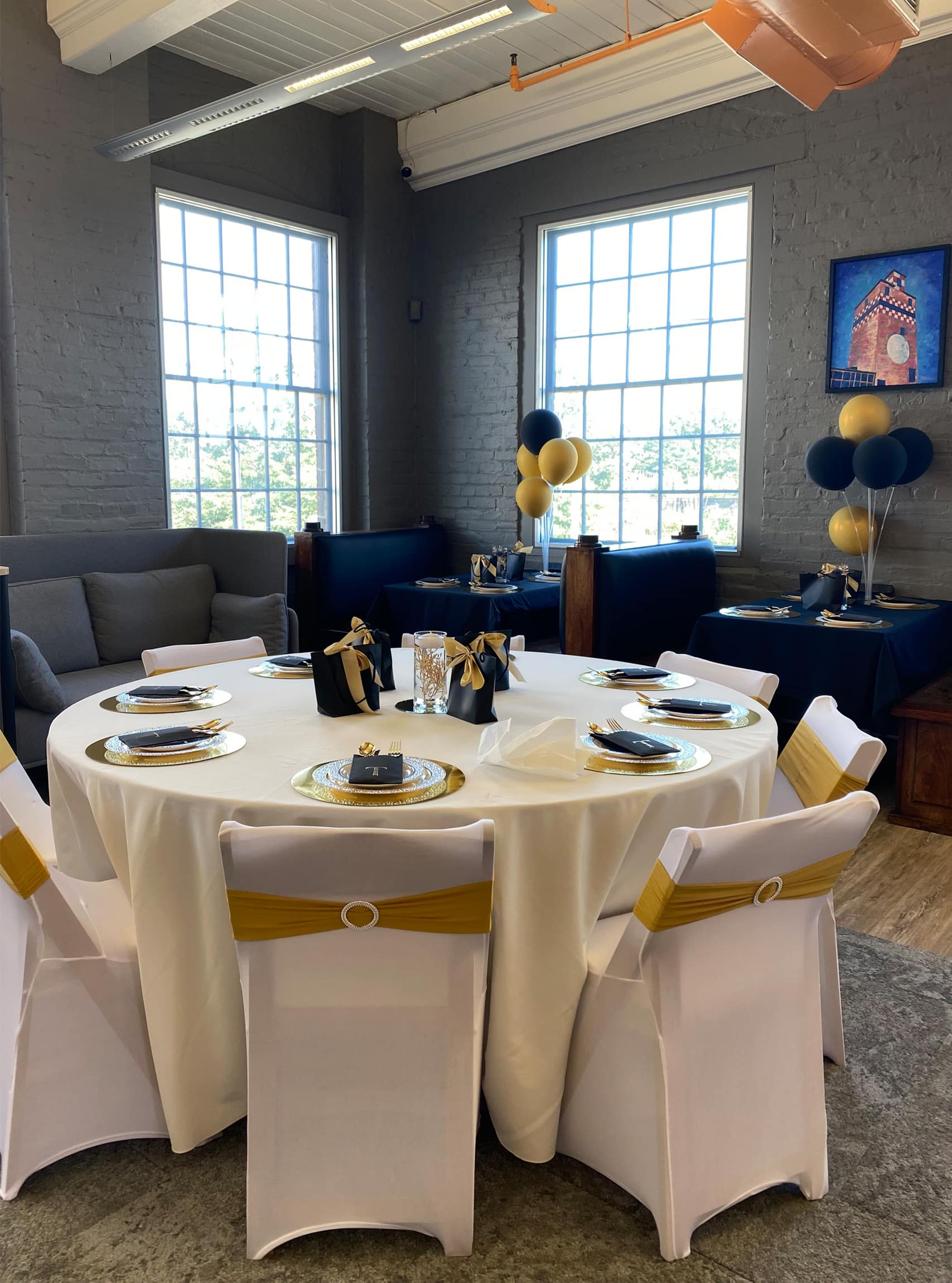 This image shows an elegantly decorated space set up for a private event at RingSpaces in Fredericksburg. The arrangement features a round table with a white tablecloth, gold accents, and neatly wrapped black party favors. Chairs are adorned with white covers and gold sashes. Additional tables with blue tablecloths and black-and-gold balloons are visible in the background. Large windows provide natural light, while the exposed brick walls and modern decor create a stylish and inviting atmosphere.