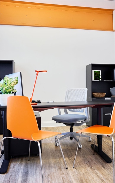 This image depicts a private office setup available at RingSpaces Coworking of Fredericksburg. The space features a modern desk, ergonomic office chair, and vibrant orange guest chairs, creating a professional and inviting environment. Minimalist decor, storage shelves, and natural light enhance the functionality and appeal, ideal for focused work or client meetings.