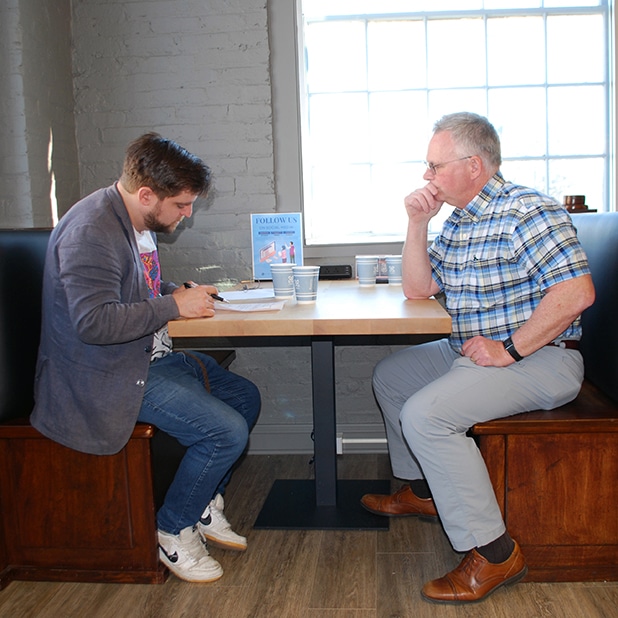 The image shows two men sitting at a table in a casual setting, likely a café or meeting space. The man on the left, dressed casually in jeans and a blazer, appears to be writing or taking notes on a document. The man on the right, wearing a plaid shirt, is seated across from him, resting his chin on his hand as if in thought or attentiveness. A small sign with the text "Follow Us" and illustrations is visible on the table, along with cups of coffee, suggesting a relaxed or informal conversation or interview. Natural light streams through the window behind them.