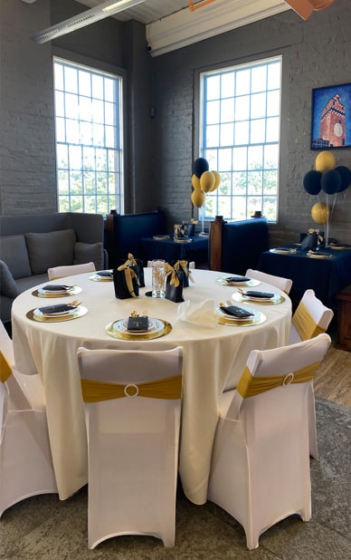 This image shows an elegantly decorated space set up for a private event at RingSpaces in Fredericksburg. The arrangement features a round table with a white tablecloth, gold accents, and neatly wrapped black party favors. Chairs are adorned with white covers and gold sashes. Additional tables with blue tablecloths and black-and-gold balloons are visible in the background. Large windows provide natural light, while the exposed brick walls and modern decor create a stylish and inviting atmosphere.