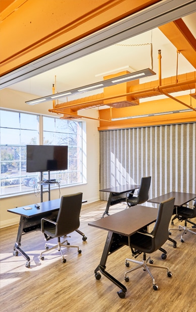 The image showcases a well-equipped meeting room available for rent at RingSpaces Coworking in Fredericksburg. The space includes multiple tables and comfortable black chairs on wheels, arranged for collaborative discussions. A large window provides natural light, while a mounted TV screen is positioned for presentations or video conferencing. The room features bright orange industrial ceiling elements and wood-style flooring, creating a modern and functional environment for professional meetings.