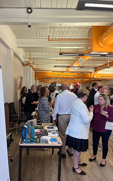 The image captures a lively business event at RingSpaces of Fredericksburg. Attendees are networking and engaging in conversations, with tables displaying books, materials, and promotional items. The open, modern coworking space, featuring bright orange ductwork and wood-style flooring, serves as a vibrant backdrop for the gathering. The event highlights RingSpaces' capability to host professional networking and business-focused occasions in a collaborative environment.