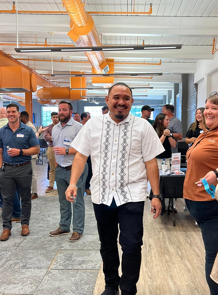 The image shows a vibrant business event at RingSpaces in Fredericksburg, with attendees smiling and engaging in a lively atmosphere. A man in traditional-style attire stands in the foreground, exuding positivity, while others mingle in the background. The space features modern elements, such as bright orange industrial accents, natural lighting from large windows, and a polished wood floor, creating an ideal venue for networking, professional events, and social gatherings.