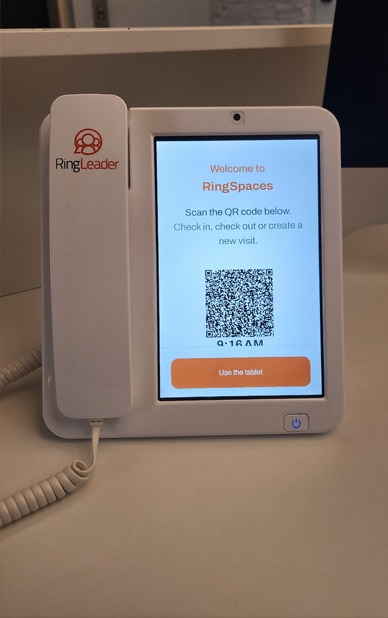 This image shows a tabletop phone system with a screen displaying the interface for "RingSpaces," a visitor management system. The screen prompts users to scan a QR code to check in, check out, or create a new visit. The device is branded "RingLeader," and the screen includes an orange button labeled "Use the tablet" for interaction.