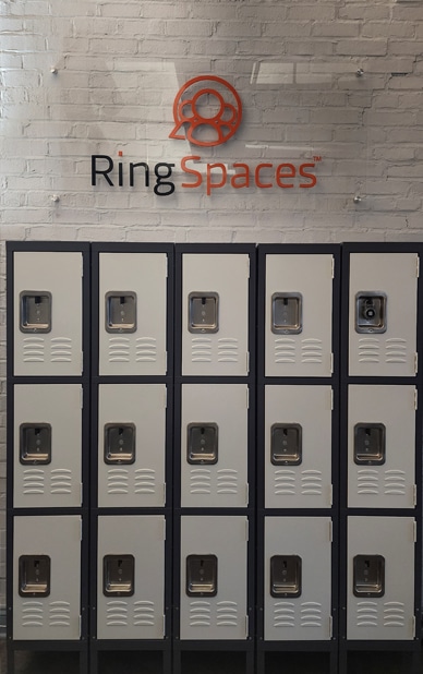 This image shows a row of secure lockers available for coworking members at RingSpaces of Fredericksburg. The lockers are neatly arranged in a grid pattern, featuring individual locks for security. The "RingSpaces" logo is prominently displayed on the wall above the lockers.