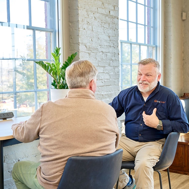 The image features a professional conversation taking place at RingSpaces Coworking in Fredericksburg. One individual, identified as Neil Darling, the owner of RingSpaces, is engaging warmly with another person. The setting reflects a collaborative and approachable environment, characteristic of the coworking space.