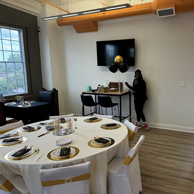 The image shows an elegantly decorated event space featuring a round table set with gold-accented tableware, white chair covers with gold sashes, and a centerpiece. In the background, there is a wall-mounted TV, balloons, and a person standing near a bar-style table. Large windows provide natural light, enhancing the modern and inviting atmosphere. This highlights the sophistication and versatility of RingSpaces in Fredericksburg as a venue for private events.