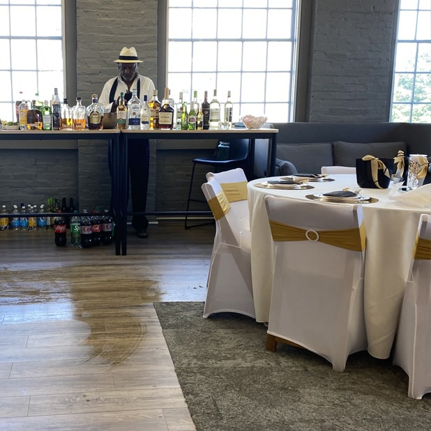 The image depicts a well-appointed event space featuring a bartender standing behind a table lined with various bottles of alcohol, with soda bottles arranged below. In the foreground, a round table is elegantly decorated with white linens, gold-accented chair sashes, and stylish place settings. The large windows allow natural light to illuminate the space, emphasizing its versatility and charm, making it ideal for private events at RingSpaces in Fredericksburg.