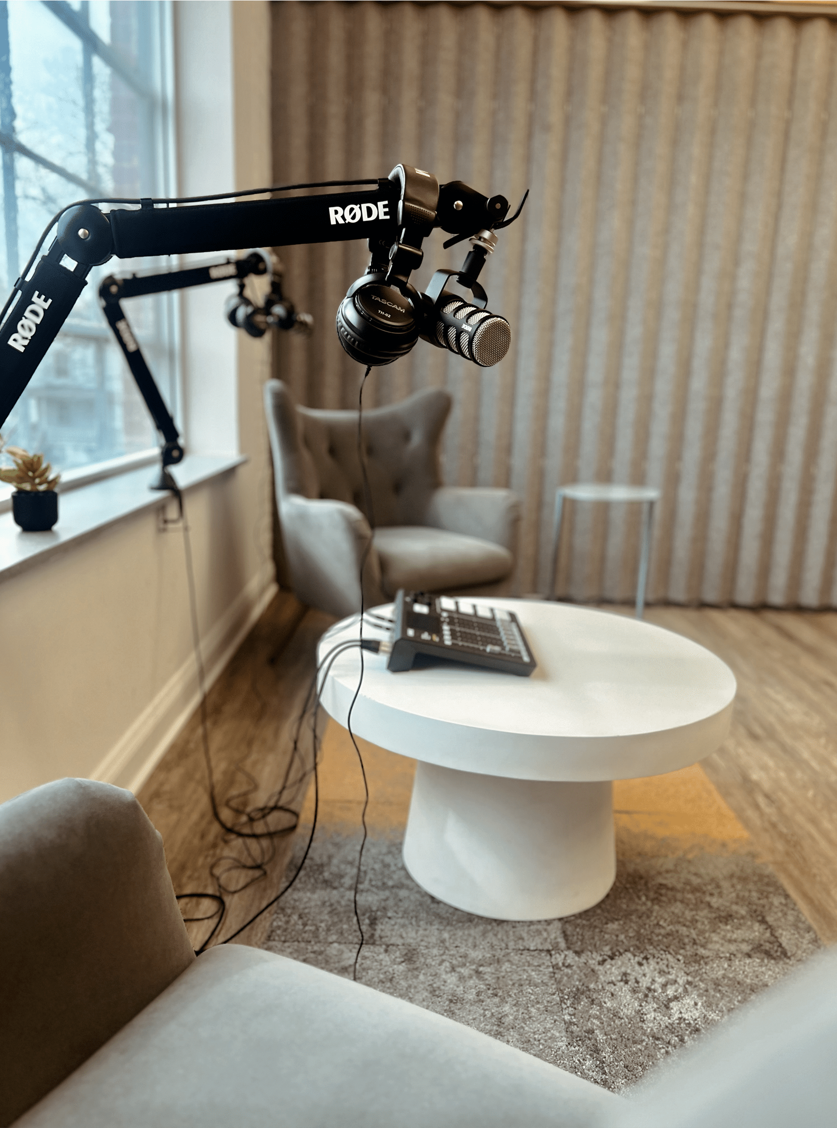 Modern podcast recording setup at RingSpaces in Fredericksburg, featuring professional microphones, headphones, a sound mixer on a round white table, and two upholstered chairs near a large window.